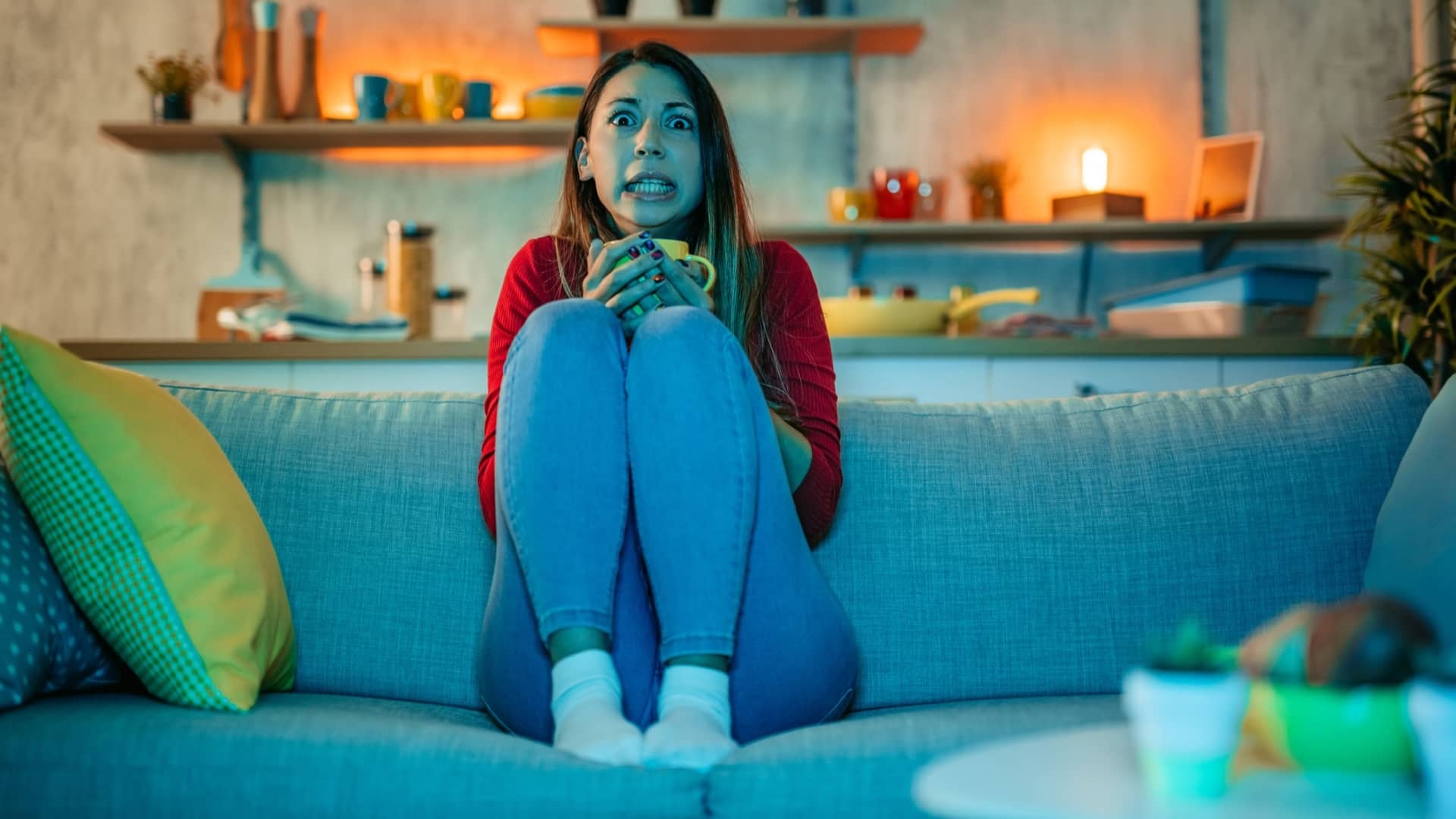 A woman watching a movie in the night and drinking coffee or tea, sitting on a couch in the living room at home, with a scared expression on her face.